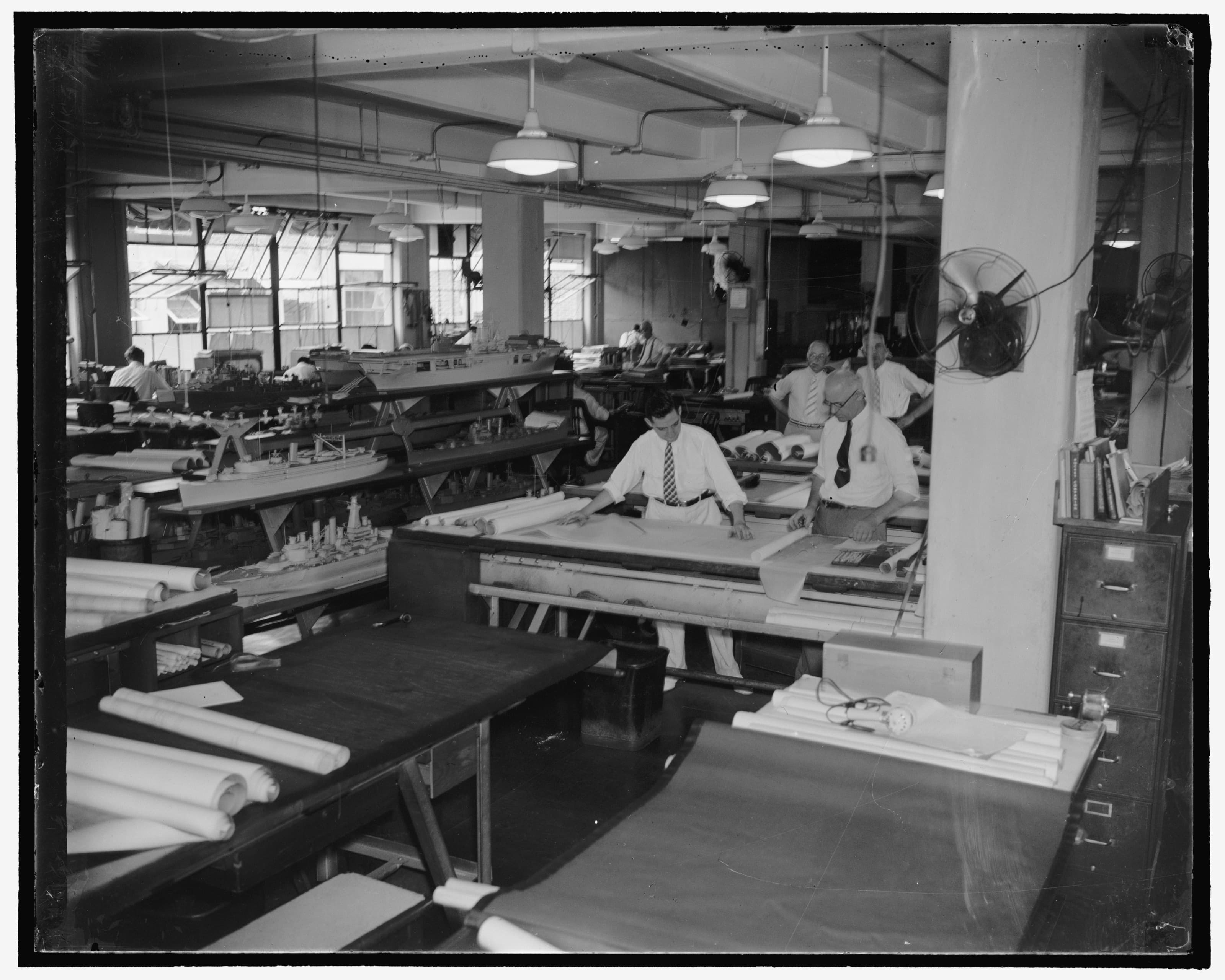 Draftsmen at work over drawing boards in the US Navy Department drafting room, rolls of drawings on every table